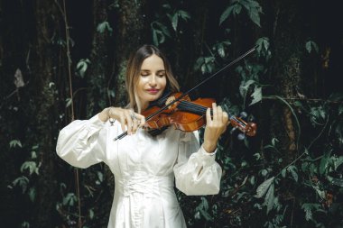 Tropikal ormanda keman çalan beyaz bir kadının portresi. Müzik, sanat konsepti. Doğada beyaz elbise giyen bir kadın. Yeşilli yeşil tropikal yaprakların arka planı. Uzayı kopyala Bali, Endonezya