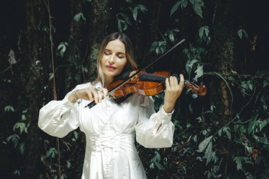 Tropikal ormanda keman çalan beyaz bir kadının portresi. Kapalı gözler. Müzik, sanat konsepti. Doğada beyaz elbise giyen bir kadın. Yeşillik tropik yaprakların arka planı. Uzayı kopyala Bali
