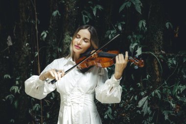 Tropikal ormanda keman çalan güzel beyaz kadın portresi. Kapalı gözler. Müzik, sanat konsepti. Beyaz elbiseli bir kadın. Yeşilli yeşil tropikal yaprakların arka planı. Uzayı kopyala Bali