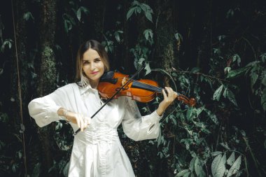 Tropikal ormanda keman çalan gülümseyen beyaz kadın portresi. Müzik, sanat konsepti. Doğada beyaz elbiseli bir kız. Yeşilli yeşil tropikal yaprakların arka planı. Uzayı kopyala Bali, Endonezya