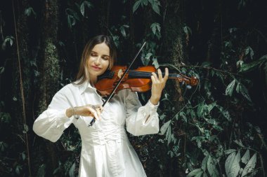 Tropikal ormanda keman çalan gülümseyen beyaz kadın portresi. Kapalı gözler. Müzik ve sanat konsepti. Beyaz elbiseli bir kadın. Yeşilli yeşil tropikal yaprakların arka planı. Uzayı kopyala Bali