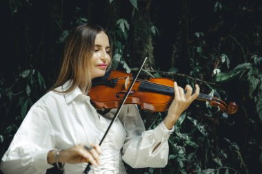 Tropikal ormanda keman çalan beyaz bir kadının portresi. Müzik, sanat konsepti. Doğada beyaz elbise giyen bir kadın. Yeşilli yeşil tropikal yaprakların arka planı. Bali, Endonezya