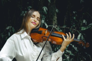 Tropikal ormanda keman çalan gülümseyen beyaz kadın portresi. Kapalı gözler. Müzik ve sanat konsepti. Sarı saçlı, beyaz elbiseli bir kadın. Bereketli yeşil tropikal yaprakların arka planı