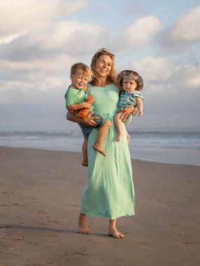 Genç anne sahilde çıplak ayakla yürüyor, kollarında iki oğul taşıyor. Aile ilişkileri. Beyaz bulutlu mavi gökyüzü. Mutlu bir çocukluk. Asya 'da yaz tatili. Seminyak Sahili, Bali, Endonezya