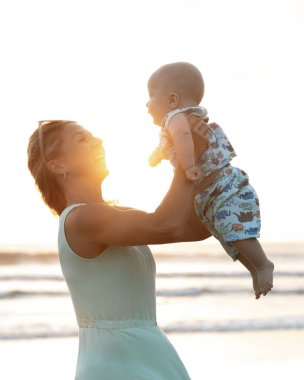 Beyaz bir anne, erkek bebeğini kucağına alıp havaya kaldırıyor. Aile dışarıda eğleniyor. Annelik kavramı. Parlak güneş ışığı. Seminyak Sahili, Bali Adası, Endonezya