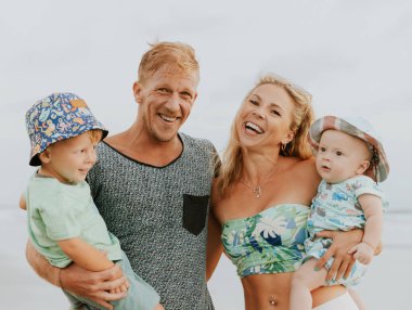 Mutlu bir ailenin sahilde vakit geçirdiği portreyi yakından çek. Anne ve baba oğullarını kucaklıyor. Sevimli erkek bebekler. Gülümseyen ebeveynler. Olumlu duygular. Asya 'da yaz tatili. Aile kavramı. Seminyak Bali