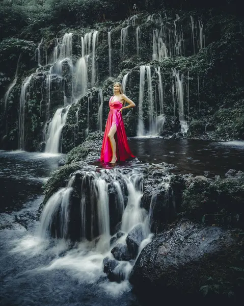 Young Caucasian woman at waterfall in tropical forest. Beautiful slim woman wearing long red dress. Banyu Wana Amertha waterfall Wanagiri, Bali, Indonesia. Slow shutter speed, motion photography