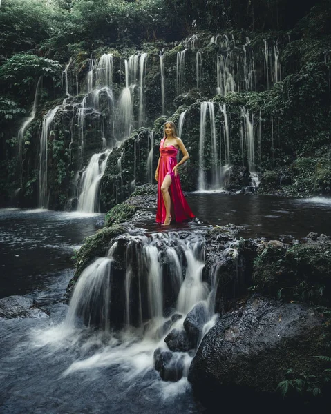 Young Caucasian woman at waterfall in tropical forest. Slim woman wearing long red dress. Beautiful pose. Banyu Wana Amertha waterfall Wanagiri, Bali, Indonesia. Slow shutter speed, motion photography