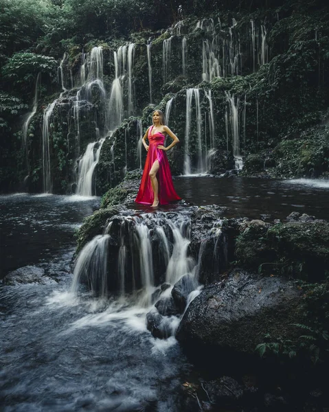 Slim Caucasian woman at waterfall in tropical forest. Young woman wearing long red dress. Beautiful pose. Banyu Wana Amertha waterfall Wanagiri, Bali, Indonesia. Slow shutter speed, motion photography