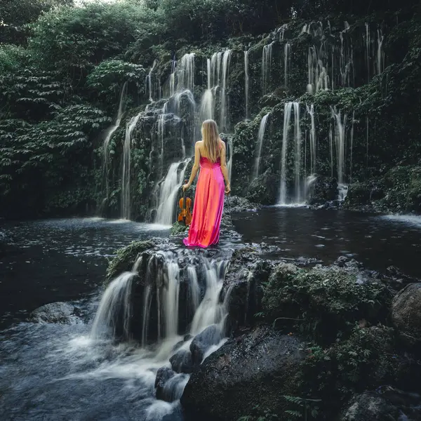 Woman with violin at waterfall in tropical forest. Music and art concept. Female wearing long red dress and holding violin in nature. Slow shutter speed, motion photography. View from back. Bali