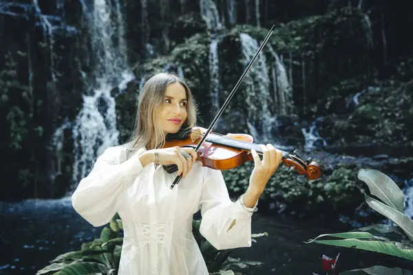 Şelale yakınlarında keman çalan güzel beyaz kadın. Müzik ve sanat konsepti. Doğada beyaz elbise giyen bir kadın. Su sıçraması. Tropik orman. Banyu Wana Amertha Şelalesi, Bali, Endonezya.