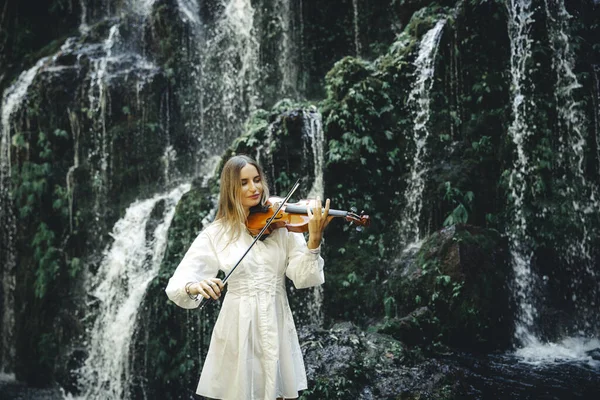 Şelale yakınlarında keman çalan beyaz bir kadın. Müzik ve sanat konsepti. Doğada beyaz elbise giyen bir kadın. Su sıçraması. Tropik orman. Banyu Wana Amertha şelalesi, Bali. Boşluğu kopyala.