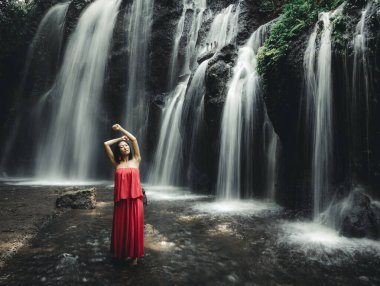 Genç Asyalı kadın şelalenin yanında poz veriyor. Doğa ve çevre kavramı. Hareket suyu. Seyahat yaşam tarzı. Uzun kırmızı elbise giyen bir kadın. İnce vücut. Uzayı kopyala Bali 'de şelale. Hareket fotoğrafçılığı.