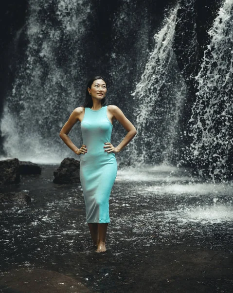 Slim Asian woman posing near the waterfall. Nature and environment concept. Travel lifestyle. Beautiful woman wearing light blue dress. Copy space. Yeh Bulan waterfall in Bali, Indonesia