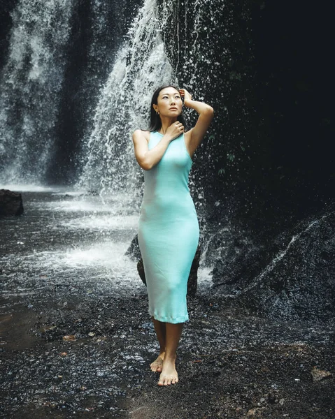 Beautiful Asian woman posing near the waterfall. Nature and environment concept. Travel lifestyle. Young woman wearing light blue dress. Slim body. Copy space. Yeh Bulan waterfall in Bali, Indonesia