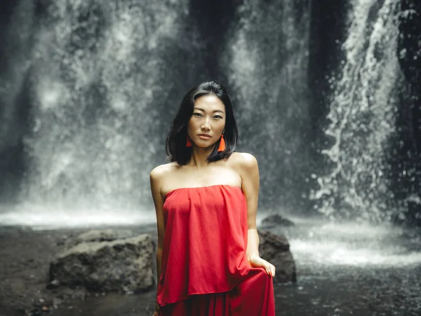 Portrait of Asian woman posing near the waterfall. Nature and environment concept. Travel lifestyle. Attractive woman wearing red dress. Copy space. Yeh Bulan waterfall in Bali, Indonesia