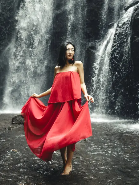 Young Asian woman posing near waterfall. Nature and environment concept. Water splashing. Travel lifestyle. Woman wearing long red dress. Slim body. Copy space. Yeh Bulan waterfall, Bali, Indonesia