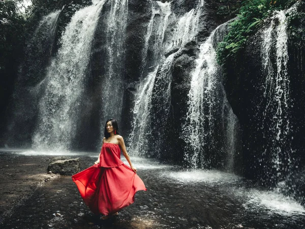 Şelalenin yanında poz veren çekici Asyalı kadın. Doğa ve çevre. Su sıçrıyor. Seyahat yaşam tarzı. Uzun kırmızı elbise giyen bir kadın. İnce vücut. Uzayı kopyala Yeh Bulan Şelalesi, Bali, Endonezya
