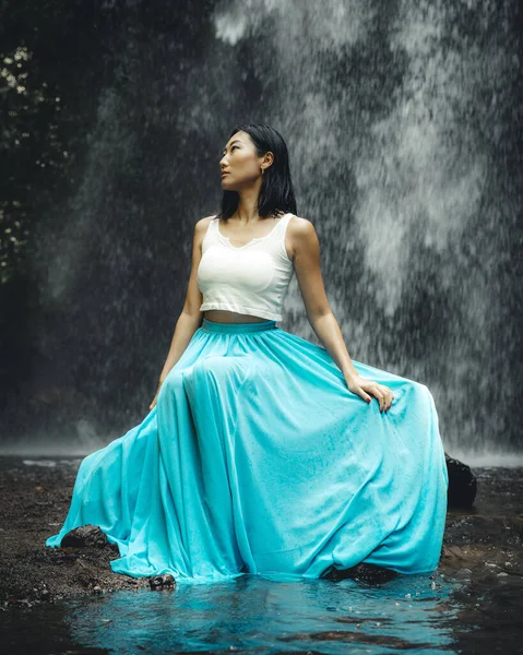 Beautiful Asian woman sitting near waterfall. Nature and environment. Water splashing. Travel lifestyle. Woman wearing long blue skirt. Slim body. Copy space. Yeh Bulan waterfall, Bali, Indonesia