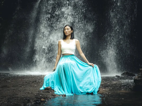 Attractive Asian woman sitting near waterfall. Nature and environment. Water splashing. Travel lifestyle. Woman wearing long blue skirt. Slim body. Copy space. Yeh Bulan waterfall, Bali, Indonesia