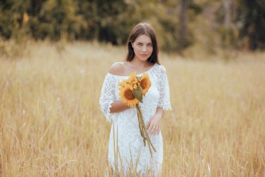 Tarlada yürüyen ve elinde bir buket ayçiçeği tutan beyaz bir kadın. Beyaz elbise giyen çekici bir kadın. Yaz tatili. Yatay yerleşim planı. Yaşam tarzı kavramı.