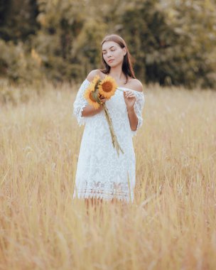 Tarlada yürüyen ve elinde bir buket ayçiçeği tutan romantik bir kadın. Beyaz elbise giyen beyaz bir beyaz kadın. Yaz zamanı. Dikey yerleşim. Yaşam tarzı kavramı.
