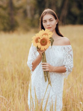 Beyaz bir kadın tarlada yürüyor ve elinde bir buket ayçiçeği tutuyor. Beyaz elbise giymiş çekici bir kadının portresi. Yaz tatili. Dikey yerleşim. Yaşam tarzı konsepti. Romantik ruh hali.