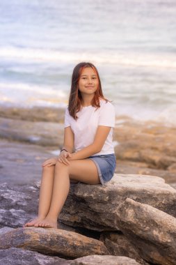 Sahildeki taşın üzerinde oturan çekici genç kız. Şort ve beyaz tişört giyen bir kadın. Asya 'da yaz tatili. Yaşam tarzı konsepti. Okyanus arka planı. Bali adasına iyi yolculuklar..