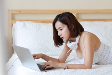 Evde bilgisayar ve kulaklık kullanırken rahat bir şekilde yatağa uzanan mutlu Asyalı kadın, yatak odası geçmişi olan güzellik işkadını. Yaşam tarzı teknoloji konsepti