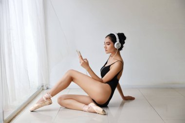Siyahi spor giyimli, kulaklıklı çekici Asyalı kadın. Cep telefonundan müzik dinleyen güzel kız bale eğitimi alırken (teknoloji ve sağlıklı konsept)