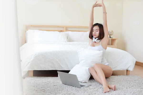 自宅でリラックスしながらパソコンやイヤフォンを使いながら 床に腰を下ろしてストレッチをするカジュアルなアジア系女性 寝室を背景にした美容室 ライフスタイル技術コンセプト — ストック写真