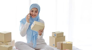 Çekici görünümlü, güzel ve Asyalı bir kadın, bilgisayar ve online paket teslimatında yatak başında oturuyor. Cep telefonuyla çalışan küçük bir işletme başlat KOBİ serbest kız 