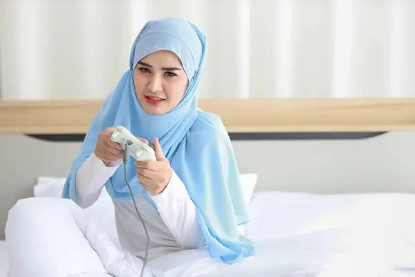 Vdo游戏控制台概念 活跃的亚洲女人穿着白色的穆斯林睡衣 坐在床上 拿着操纵杆 玩着令人兴奋的游戏 可爱的女孩对游戏控制器感到兴奋 — 图库照片