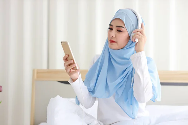 穿着穆斯林睡衣的美丽的亚洲女人的画像 在手机上观看在线故事 坐在床上 与无线网络相连 年轻可爱的戴头巾的女孩听智能手机里的音乐 — 图库照片