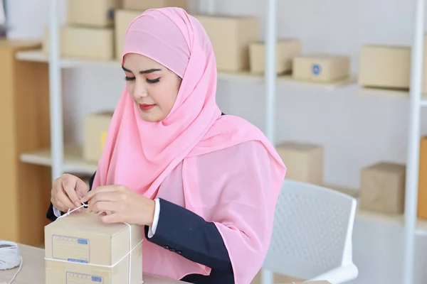 积极的笑着的亚洲女人穿着蓝色的穆斯林西服坐着 与网上包裹箱递送一起工作 创业的中小企业自由职业女孩 在电脑和手机上工作 面带微笑 — 图库照片