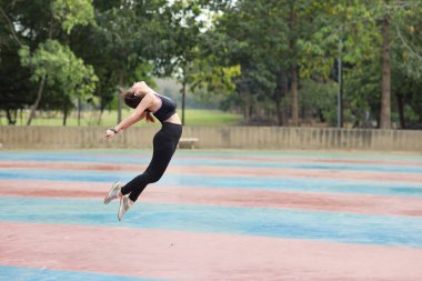 Spor giyimli genç Asyalı atletik kadın ayakta duruyor ve sabah egzersizi için dışarıda kardiyo antrenmanı yapıyor. Yeşil ağaçtan atlayan aktif ve sağlıklı bir kız. Spor konsepti