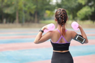 Dikiz ayarı atletik genç Asyalı kadın spor kıyafetleriyle ayakta duruyor ve sabah egzersizi için dambılları kaldırıyor. Aktif kız, ağaç köklü pazı egzersizi yapıyor. Spor konsepti