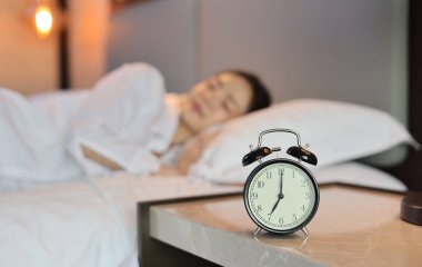 Üzerinde uyuyan, beyaz uyku tulumu giymiş, yatakta uzanmış, mutluluk ve gülümsemeyle uyuyan bir kadının bulanık görüntüsü olan çalar saat. Yaşam tarzı kavramı