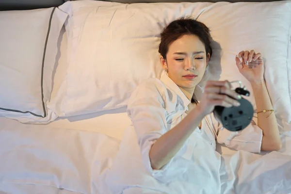Happy young asian woman in white lingerie lying in bed, wake up late and overslept morning, trying to stop alarm clock. Cute girl look comfortable, need more sleep. Difficult morning get up concept