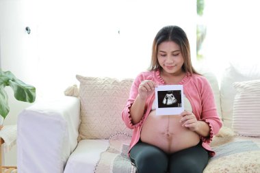 Mutlu gülümseyen Asyalı hamile kadın ultrason görüntüsü gösterirken oturma odasında dinleniyor ve koltukta oturuyor. Müstakbel anne hamilelik süresince doğum için hazırlanıyor ve bekliyor..