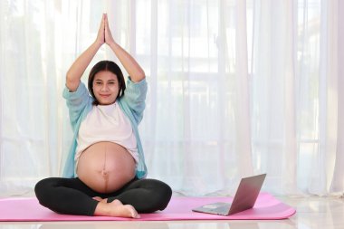 Mutlu Asyalı hamile, yoga minderinde oturup yoga yapıyor ve evdeki bilgisayardan eğitim videosu izliyor. Müstakbel anne hamilelik konsepti sırasında bebek doğurmaya hazırlanıyor.