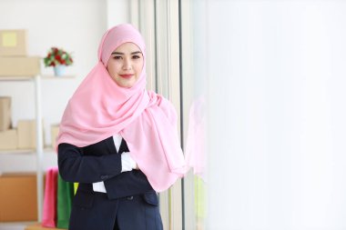 Mavi takım elbiseli, pembe şaftlı, dindar, Asyalı Müslüman kadın ayakta kameraya özgüvenle bakıyor. İş kadını, paket KOBİ kutu teslimat geçmişiyle ayakta duruyor. Ev konseptinde çalış