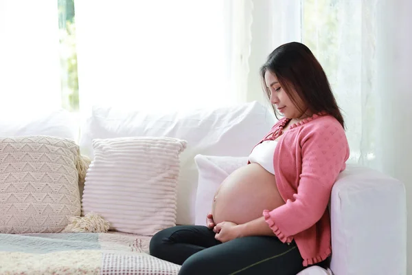 Happy smiling young asian pregnant woman sitting and resting on sofa in living room while touching and looking her belly. Expectant mother preparing and waiting for baby birth during pregnancy.