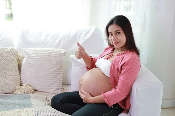 Happy smiling young asian pregnant woman resting and sitting on sofa in living room while touching and showing thumb. Expectant mother preparing and waiting for baby birth during pregnancy.