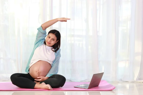 Happy asian pregnant sit on yoga mat practice stretching yoga and watch training video online from computer in living room at home. Expectant mother prepare for baby birth during pregnancy concept