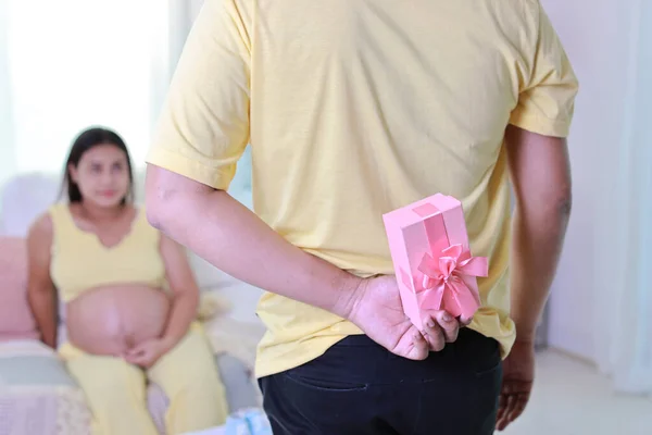 Happy asian pregnancy sitting and resting on sofa in living room while husband standing and hiding surprise gift. Expectant mother preparing and waiting for baby birth during pregnancy.