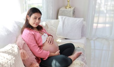 Mutlu gülümseyen Asyalı hamile kadın oturma odasında koltukta oturup dinlenirken karnına ve kamerasına dokunuyor. Müstakbel anne hamilelik süresince doğum için hazırlanıyor ve bekliyor..