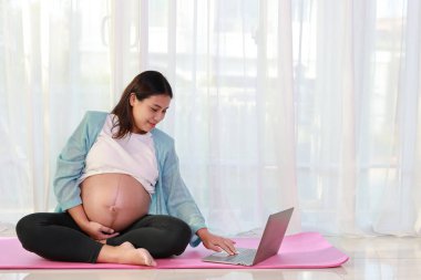 Mutlu Asyalı hamile, yoga minderinde oturup yoga antrenmanı yapıyor ve evdeki bilgisayardan eğitim videosu izliyor. Müstakbel anne hamilelik konsepti sırasında bebek doğurmaya hazırlanıyor.