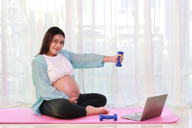 Mutlu genç Asyalı hamile, pembe paspasın üzerine oturmuş halteri kaldırıyor ve evdeki bilgisayardan eğitim videosu izliyor. Müstakbel anne hamilelik konsepti sırasında bebek doğurmaya hazırlanıyor.