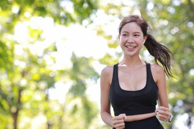 Güzel Asyalı fitness bayan sporcu. Sabah koşusu antrenmanı yapıyor. Açık havada. Parkta ağaçların altında koşan neşeli gülümseyen atlet kadın kameraya bakıyor ve müzik dinliyor.